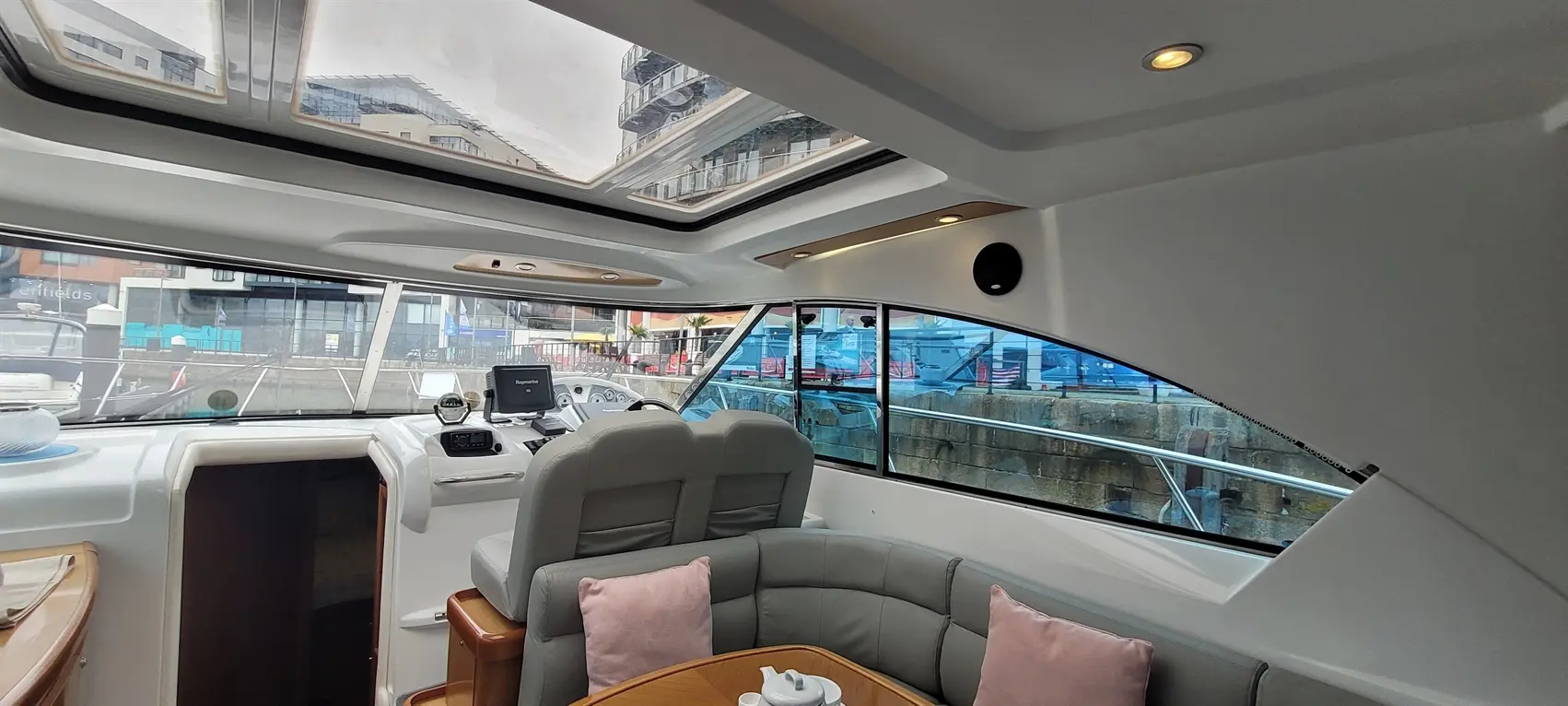 Interior of a yacht featuring a steering console, grey seating, skylights, and a view of the marina outside with buildings.